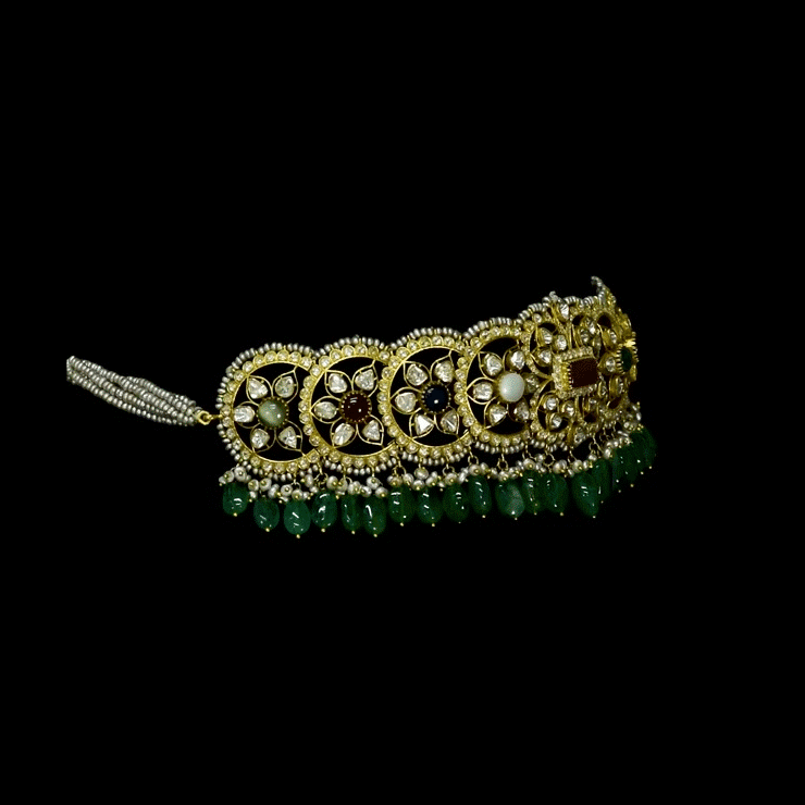 A fusion Navratan choker featuring polki diamonds in gold, highlighted with a central ruby, multicolored gemstones, intricate pearl detailing, and cascading emerald drops—paired with statement earrings for a rich, regal presence. (KMNE3719)